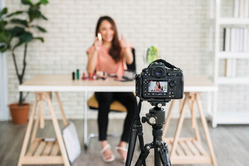 Young Asian beautiful woman professional beauty vlogger or blogger sell lipstick and cosmetic products to share on online social media. Focus at Live video screen of dslr camera by tripod.