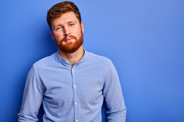 intelligent guy in shirt posing at camera isolated over blue background, handsome red haired male with beard