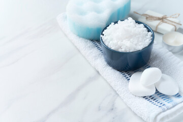 Spa resort therapy composition with dark blue bowl full bath salt, white stones, soap, bath sponge on a towel on the marble background. Copy space