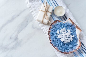 Fototapeta na wymiar Beautiful spa composition with big sea shell full of blue and white bath salt, soap, candle on a towel on the light marble background. Copy space. Flat lay