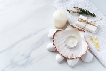 Spa supplies on the light marble background. Big sea shell with water and candle, white stones, soap, vials of oil, towel and candle. Copy space. Flat lay