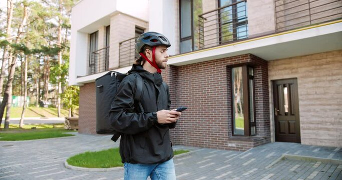 Portrait of young male courier in helmet with big backpack walking on street and texting on smartphone. Caucasian delivery man typing on cellphone searching for address. Delivery service concept