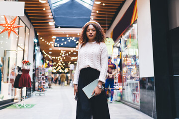 Half length portrait of trendy dressed african american female standing in shopping mall during free time, attractive 20s dark skinned woman dressed in stylish outfit posing in store looking at camera