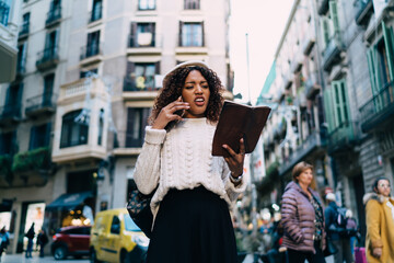 Confused black woman talking on smartphone and reading book on street