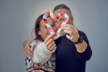 Incognito adult couple in Christmas love concept, holding paper cut candy canes in a shape of a heart in their hands, standing close and embracing. Blurred portrait against grey background in studio