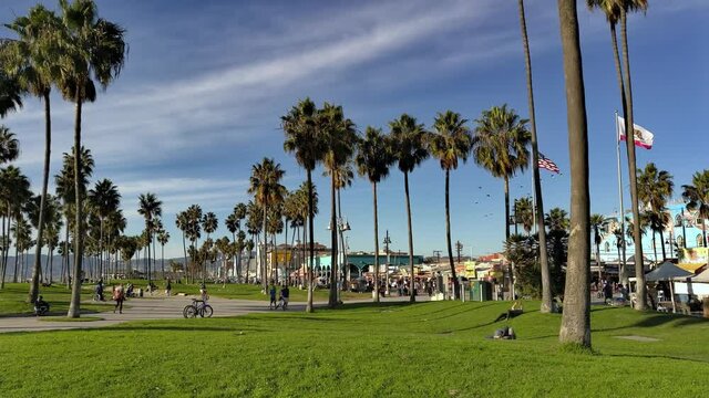 People walking around the Venice Beach boardwalk in the afternoon with palm trees and the American flag flying in the background in Los Angeles, Califonia, USA - Wide Shot