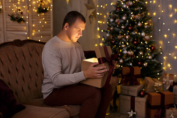 Christmas and magic concept - portrait of young man opening gift box in decorated room with Christmas tree and led lights