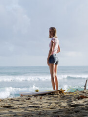 Thoughtful woman stands on the beach, a surfboard lying on the sand.