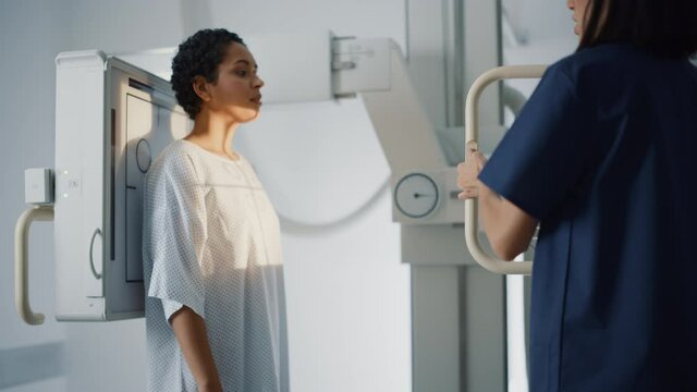 Hospital Radiology Room: Beautiful Latin Woman Standing while Professional Female Radiologist Explains Procedure, Adjusts X-Ray Machine