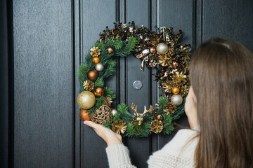 Young woman hanging christmas festive wreath on house door. Traditional home decoration on winter...