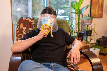 Medium close shot of a young man relaxing in an armchair indoor and eating a banana, wearing protective face mask and anti covid plastic visor.