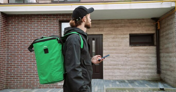 Portrait of Caucasian male worker with big green thermal bag walk on street typing on smartphone looking for address in navigation app to deliver order. Delivery man an typing on cellphone. Side view