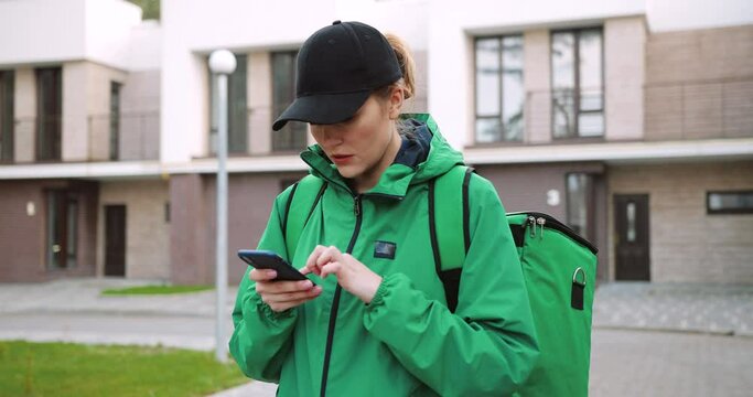 Portrait of young Caucasian female worker in green jacket with big thermal bag standing on street typing on smartphone looking for address in navigation app to deliver order. Woman typing on cellphone