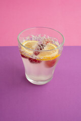 a glass of sparkling water cocktail with sliced lemon, cherry and berry. non-alcoholic drink, beverage. purple textured backdrop and pink background. carbonated water, bubble. abstract close up.