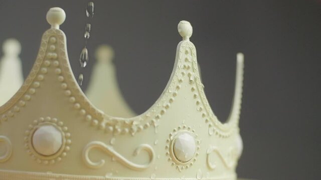 Close up of a beige crown with ornaments and white pearl jewels with peaks jagged out of plastic with water drops dripping on it. Smooth sliding camera movement. Shot in 4K.	