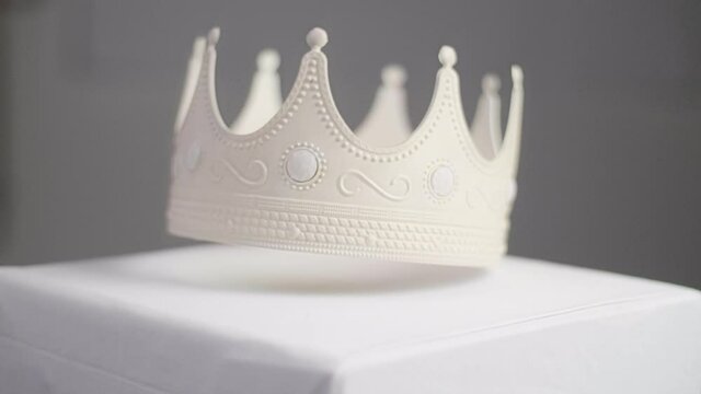 Beige crown with ornaments and white pearl jewels with peaks jagged out of plastic falling on a white pedestal; slow motion. With a neutral grey background with shallow depth of field. Shot in 4K.	