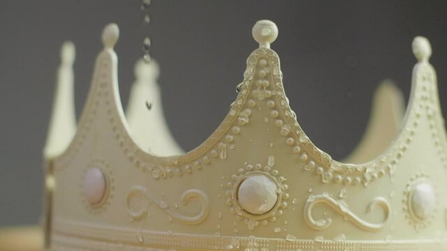 Close up of a beige crown on a pedestal with ornaments and white pearl jewels with peaks jagged out of plastic with water drops dripping on it. Smooth sliding camera movement. Shot in 4K.	
