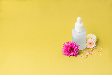 Beauty product package on yellow background, pink flowers and sea salt sprinkled. Blank white serum bottle with pipette and no branding. Place text