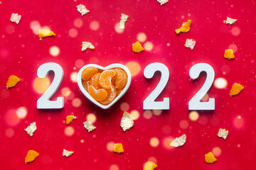 on a red background white numbers 2022. instead of the number 0, a plate in the shape of a heart with slices of tangerines. tangerine peel scattered like confetti