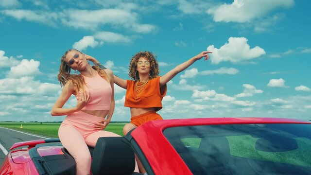 Portrait of two young beautiful hipster girls in red convertible car. Sexy carefree women in a cabriolet. Positive models is riding and having fun. Raising hands at summer and blue sky on background.