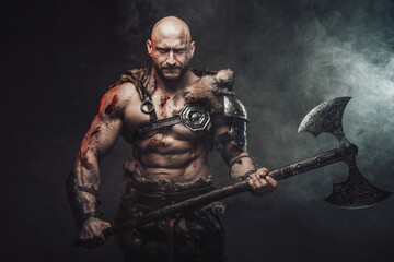 Fototapeta na wymiar Atmospheric portrait of a powerful and hairless scandinavian warrior posing in dark and smokey background holding his steel two handed axe looking at camera.