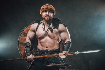 Fototapeta na wymiar Muscular and savage viking with naked torso and shield on his back poses holding a spear and looking at camera in dark and smokey background.