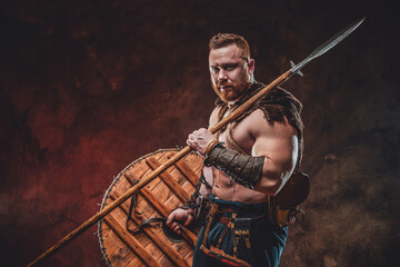 Nordic warlike barbarian with muscular build and naked torso holding shield and spear in dark...