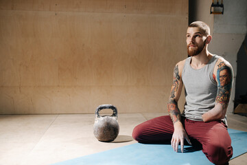 Serene disabled armless man training, sitting on his legs on a mat next to a kettlebell