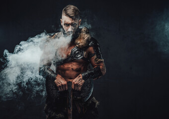 Brutal nordic heathen dressed in light armour posing in dark smokey background with huge axe and staring at camera.