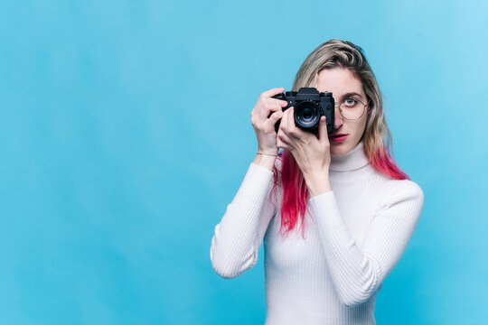 Caucasian girl with blonde and pink hair takes a picture with a camera isolated on a blue background