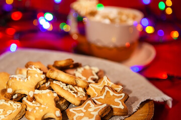 Christmas gingerbread cookies on a wooden stand and a cup of hot chocolate with marshmallows in the back, on a red background with a beautiful bokeh of glowing garland. Christmas and New Year concept.