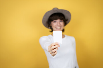 Young caucasian woman wearing hat over isolated yellow background smiling and holding white card