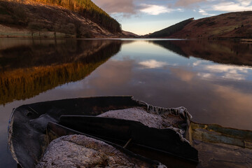 Winter morning sky reflected in lake with a frosty small boat in the foreground. Llyn Geirionydd, Trefriw, Snowdonia. Taken soon after sunrise in January.