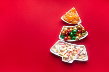 Obraz na płótnie Canvas on a red background there is a tray in the form of a Christmas tree. sweets in the tray: marshmallows, round candies in foil and tangerine slices
