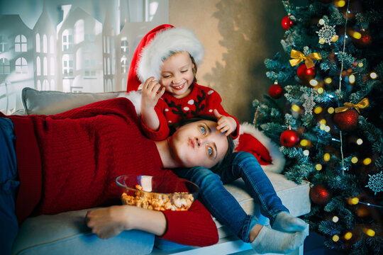 A young mother and her daughter in Santa hats watch movies at home for Christmas and eat popcorn