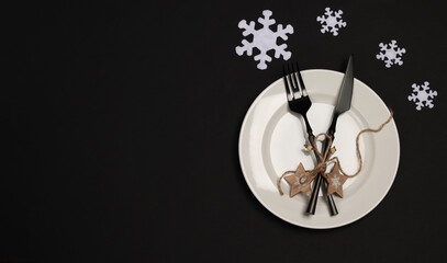 Christmas and New Year table setting. White plate, snowflakes, black cutlery on a black background. Modern and elegant. Copy space. flat lay