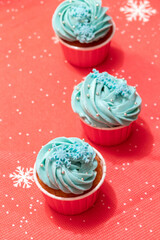 Obraz na płótnie Canvas Cupcakes with blue cream, snowflakes and silver beads on red tablecloth. Christmas dessert. Selective focus.