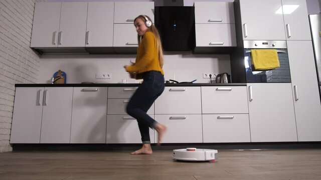 Happy pretty female in headphones listening to music and dancing while robotic vacuum cleaner doing dirty work. Young woman enjoying free time while automatic hoover cleaning floor in home kitchen