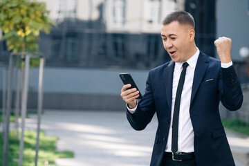 Fototapeta premium Portrait of excited man reading good news at smartphone outdoors. Successful businessman celebrating win with mobile phone outside. Surprised man looking mobile phone outside