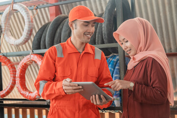 male mechanic in a wearpack uniform and a female customer using a digital tablet to view the catalog at a spare part workshop