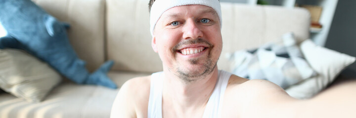 Obraz premium Joyful young man in sport headband holding pink dumbbell and taking picture with smartphone