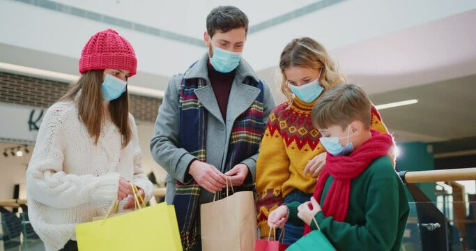 Caucasian Happy Family Wearing Masks Visiting Shopping Mall During Quarantine And Showing Their Purchases In The Bags. Cute Children. Parenthood. Christmas Tradition.