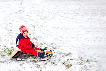 Fototapeta na wymiar Little boy in knitted striped hat and warm red jacket enjoy sledding at park on Christmas holidays.Cute baby wearing warm clothes play with snow on back yard near home. Outdoor fun for family vacation