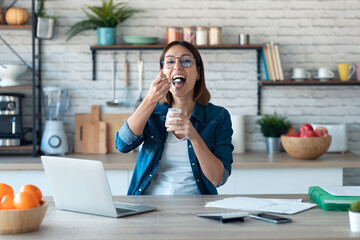 Beautiful young business woman working with computer while eating yogurt in the kitchen at home.