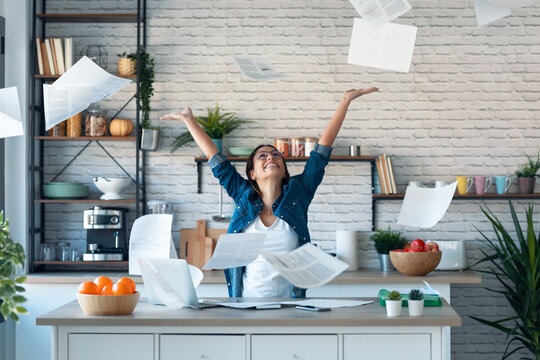 Happy young woman tossing invoices and documents into the air while working on the laptop at home.