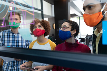 Diverse group of colleagues wearing face masks brainstorming at glass wall in office