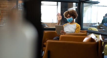 Mixed race woman wearing face mask using tablet and earphones waving during video meeting