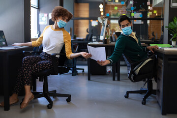 Mixed race woman and man wearing face masks passing document in casual office