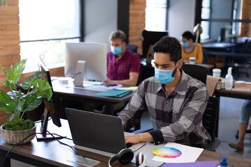 Diverse male colleagues wearing face masks working in computers at office desks