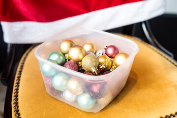 Golden, tiffany and burgundy Christmas balls in a transparent plastic food container on vintage orange chair with red cover. Storage of new year decoration. Space management